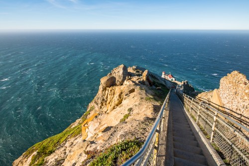 A winding staircase leads down a rocky cliff to a lighthouse overlooking a vast ocean under a clear blue sky.