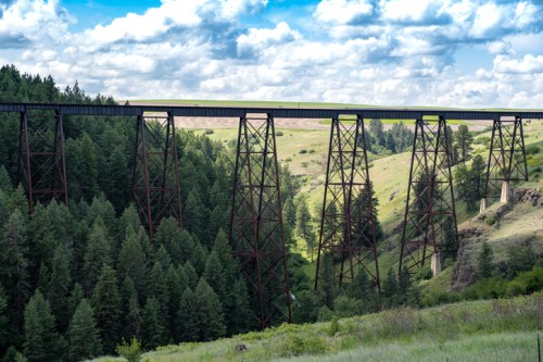 A tall, metal trestle bridge spans a lush green valley, surrounded by dense forests and a blue sky with fluffy clouds.