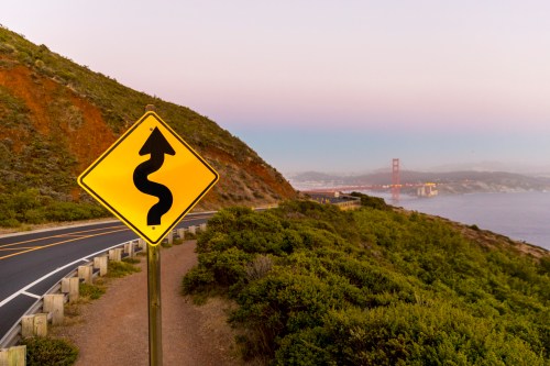 A winding road sign near a coastal path, with the Golden Gate Bridge visible in the background at dusk.