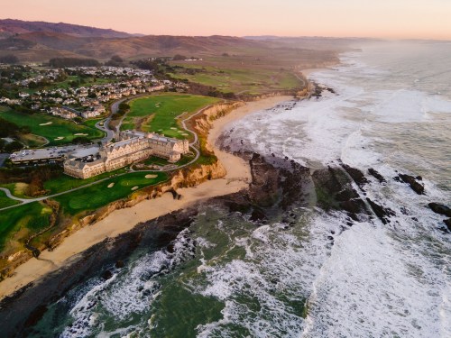 Aerial view of a coastal resort near a golf course, with waves crashing on rocky shores at sunset.