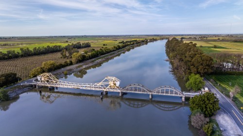 Aerial view of a white bridge over a calm river, surrounded by green fields and trees under a clear blue sky.