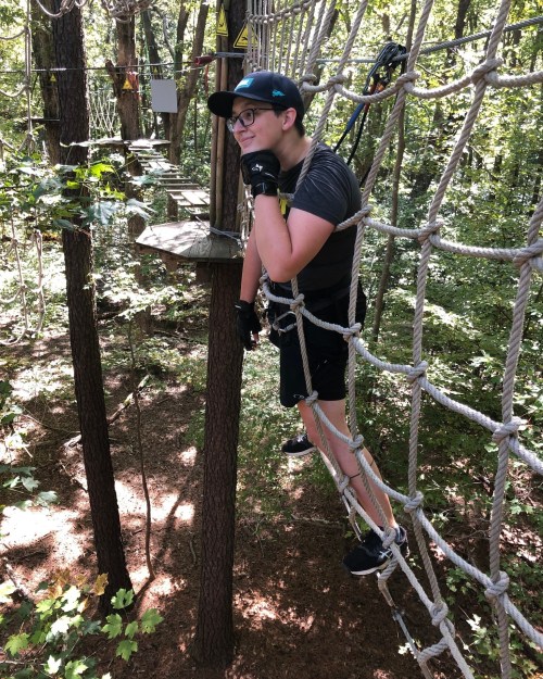 A person in a black shirt and cap balances on a rope course among trees, looking thoughtfully to the side.