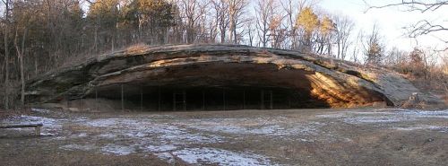 A large rock overhang in a wooded area, with a grassy foreground and patches of snow. Sunlight illuminates the rock face.