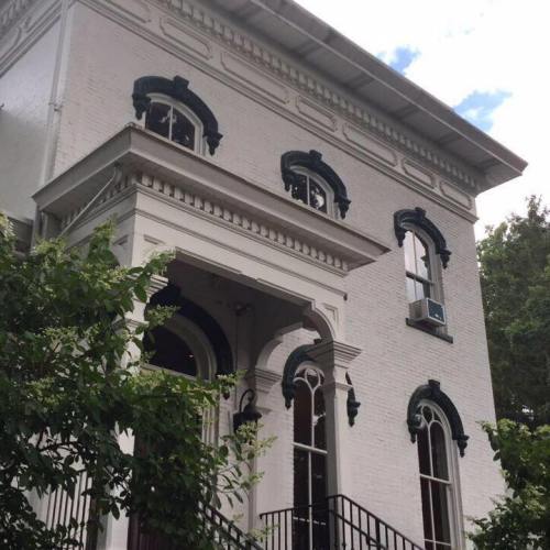 A historic white brick building with decorative blue trim and large windows, surrounded by greenery.