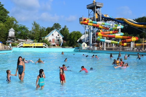 A lively water park scene with children and adults swimming, playing, and enjoying colorful water slides under a blue sky.
