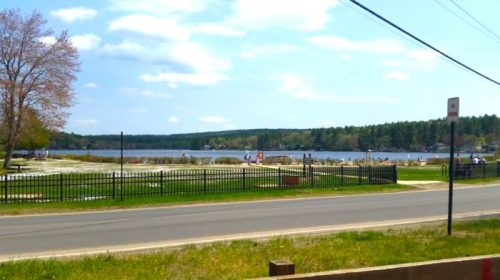 A scenic view of a lake surrounded by trees, with a grassy area and a fence in the foreground under a blue sky.