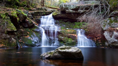 A serene waterfall cascades over moss-covered rocks into a calm pool, surrounded by autumn foliage.