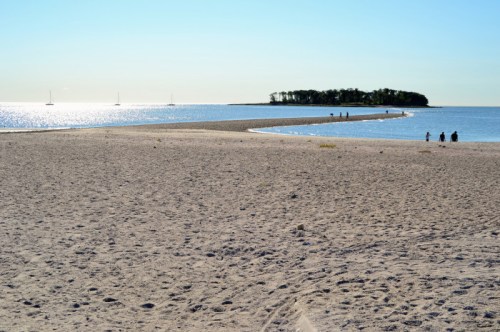 A sandy beach extends into calm waters, with a small island in the distance and a few people walking along the shore.