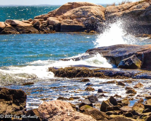 Waves crashing against rocky shoreline with blue water and distant land under a clear sky.