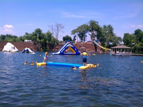 A sunny lake scene with children swimming and playing on inflatable structures, surrounded by trees and a gazebo.