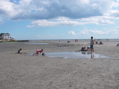 Children playing in shallow water on a sandy beach, with people relaxing in the background under a partly cloudy sky.