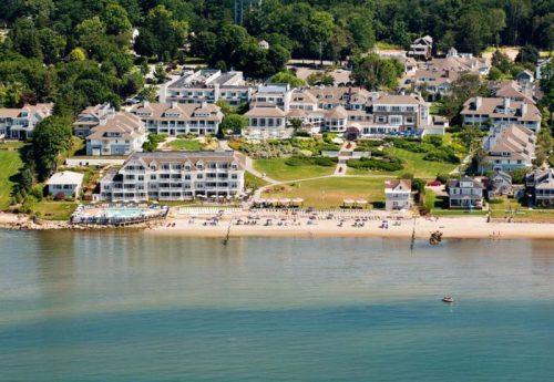 Aerial view of a beach resort with white buildings, green lawns, and a sandy beach along a calm water body.