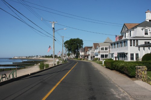 A coastal road lined with houses and American flags, leading to a calm beach and clear blue sky.