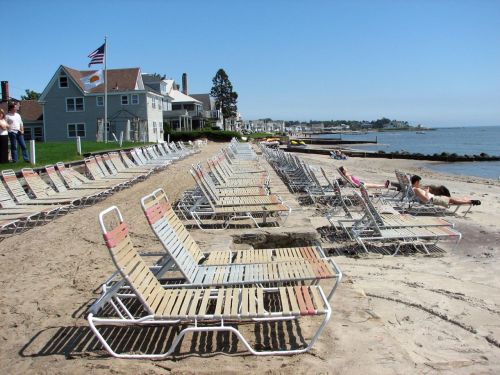A sandy beach lined with empty lounge chairs, with a few people relaxing in the sun and houses in the background.