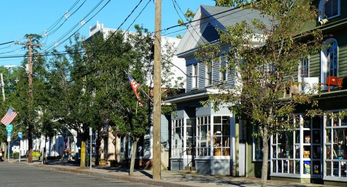 A charming street lined with trees and quaint shops, featuring American flags and a clear blue sky.
