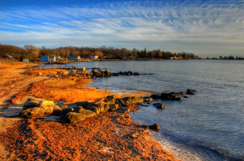 A serene shoreline with rocks and golden grass, under a blue sky with wispy clouds reflecting on calm water.