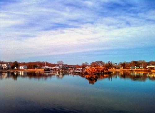 A serene waterfront scene with calm water reflecting houses and trees under a blue sky with wispy clouds.