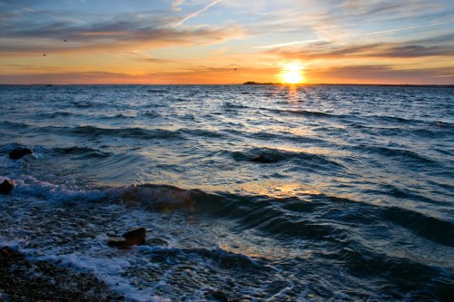 Sunset over a calm ocean, with gentle waves lapping at a rocky shore under a colorful sky.