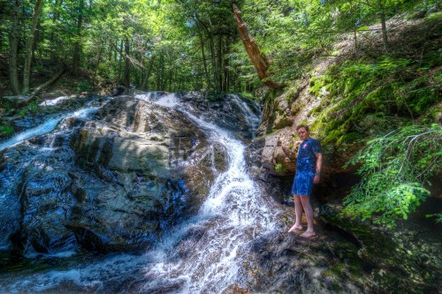 A person stands barefoot by a cascading waterfall in a lush, green forest. Sunlight filters through the trees.