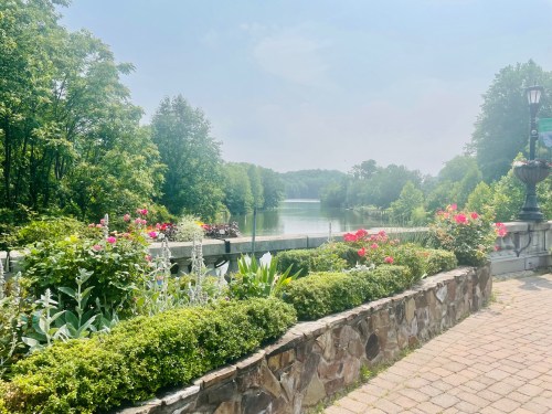 A serene view of a river surrounded by lush greenery and colorful flowers, with a stone pathway in the foreground.