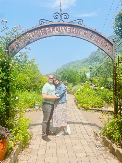 A couple stands under a decorative archway in a lush garden, smiling and embracing on a sunny day.