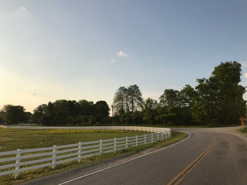 A winding road curves alongside a white fence and green trees under a clear blue sky.