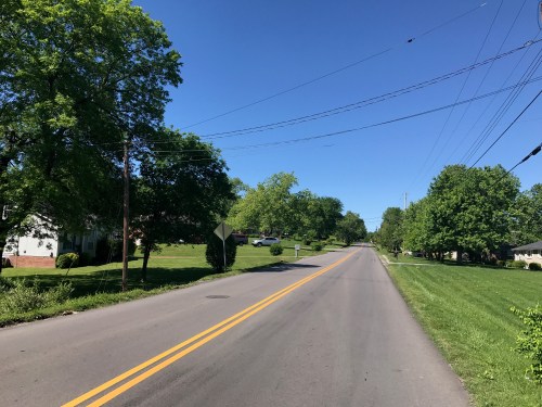A quiet, tree-lined road under a clear blue sky, with green lawns and houses visible in the distance.