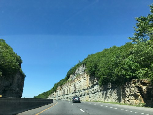 A car drives on a highway surrounded by tall, rocky cliffs and lush green trees under a clear blue sky.
