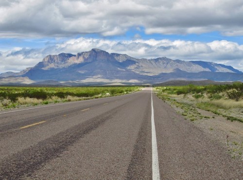 A long, empty road stretches toward a distant mountain range under a partly cloudy sky. Green fields line the sides.