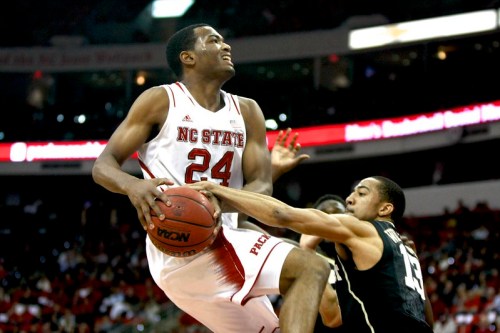 A basketball player in a red NC State uniform leaps for a shot while being defended by an opponent in a black uniform.