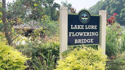 Sign for Lake Lure Flowering Bridge surrounded by greenery and flowers, with a gazebo visible in the background.