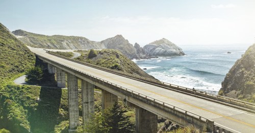 A scenic coastal highway curves along cliffs, overlooking the ocean and rocky formations under a clear sky.
