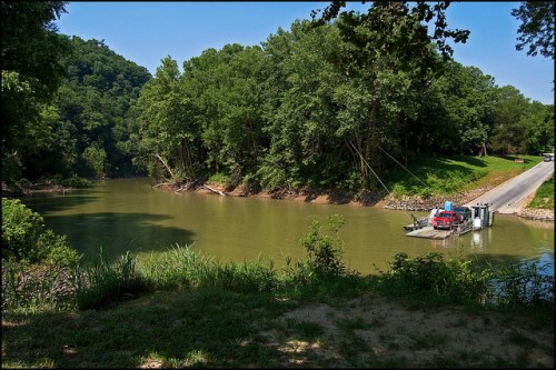 A serene river scene with lush greenery, a ferry on the water, and a road lined with trees in the background.