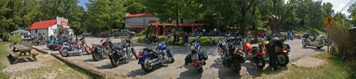 A panoramic view of a motorcycle gathering outside a rustic venue, with numerous bikes parked and people socializing.