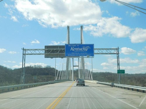 A road leading to a bridge with a "Welcome to Kentucky" sign against a blue sky with clouds.