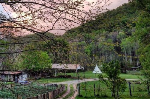 A serene farm scene with a barn, a teepee, and lush greenery under a colorful sky at sunset.