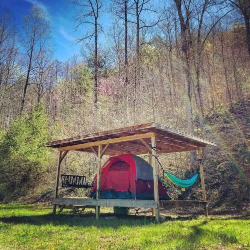 A red tent under a wooden shelter with a hammock and swing, surrounded by trees on a sunny day.