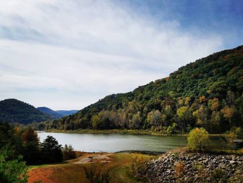Scenic view of a calm lake surrounded by lush, colorful hills under a partly cloudy sky.