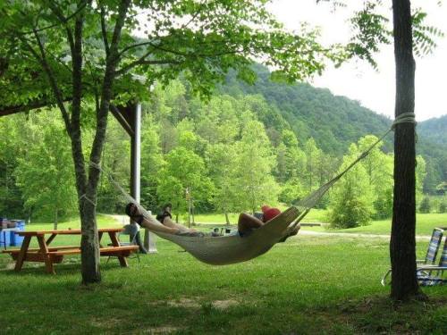 A person relaxes in a hammock between two trees, surrounded by green grass and mountains in the background.