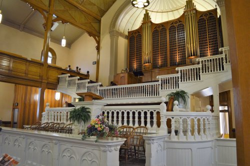 Interior of a church featuring a large organ, wooden pews, and decorative railings, with people seated in the balcony.