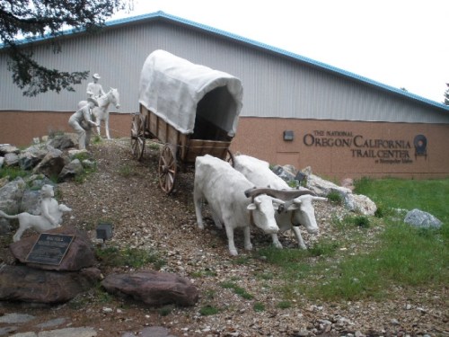 A covered wagon pulled by two white oxen, with a cowboy figure, in front of the Oregon-California Trail Center.