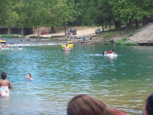 A scenic swimming area with people enjoying the water, surrounded by trees and a sandy beach in the background.
