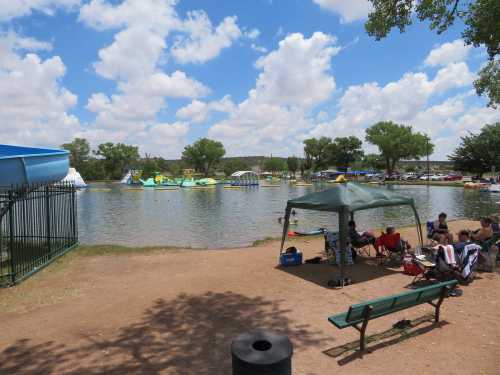 A sunny park scene with a lake, inflatable water attractions, and people relaxing under a canopy.