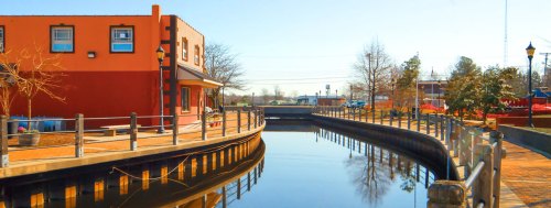 A peaceful canal scene with a colorful building, trees, and a walkway along the water under a clear blue sky.
