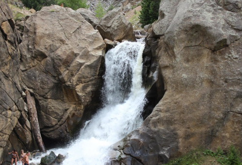 A waterfall cascades over large rocks, surrounded by greenery and rugged terrain.