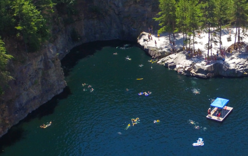 Aerial view of a swimming area with people in the water and a floating dock surrounded by rocky cliffs and trees.
