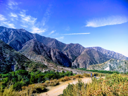 A scenic view of mountains under a blue sky, with a dirt path and two people walking in the foreground.