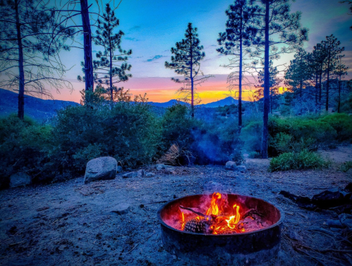 A campfire burns in a metal ring, surrounded by trees and mountains under a colorful sunset sky.