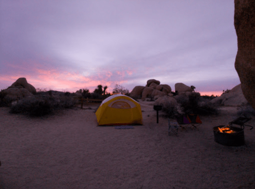 A yellow tent in a desert campsite at dusk, with rocks and a fire pit nearby under a colorful sky.