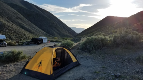 A yellow tent set up in a mountainous campsite, with hills and a sunset in the background.